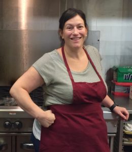 a woman in a maroon apron leaning against an industrial oven